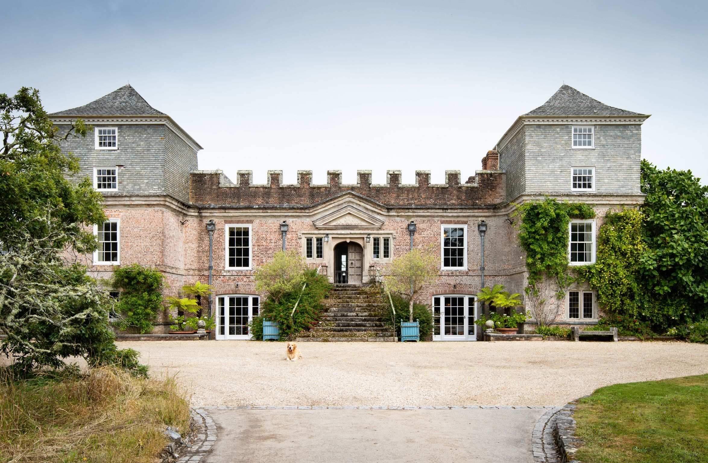 The north front of Ince Castle: a rose-brick house with battlements and a slate-hung tower at each corner, behind a gravel forecourt